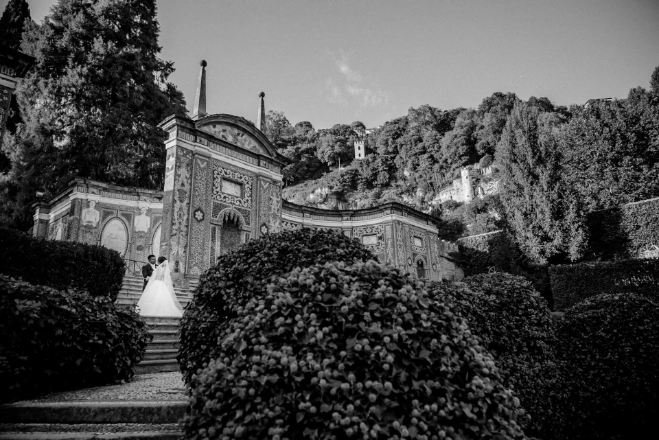 A bride and groom on garden steps beside an ornate facade, in black and white.