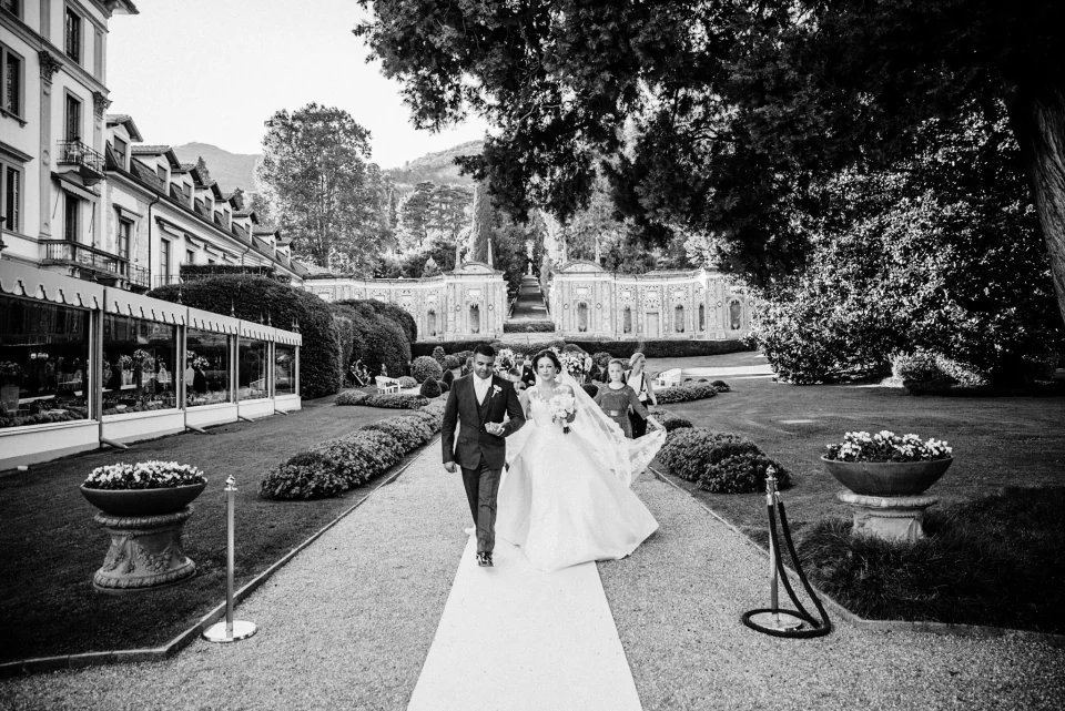 A bride and groom walking through formal gardens with their wedding guests behind them.
