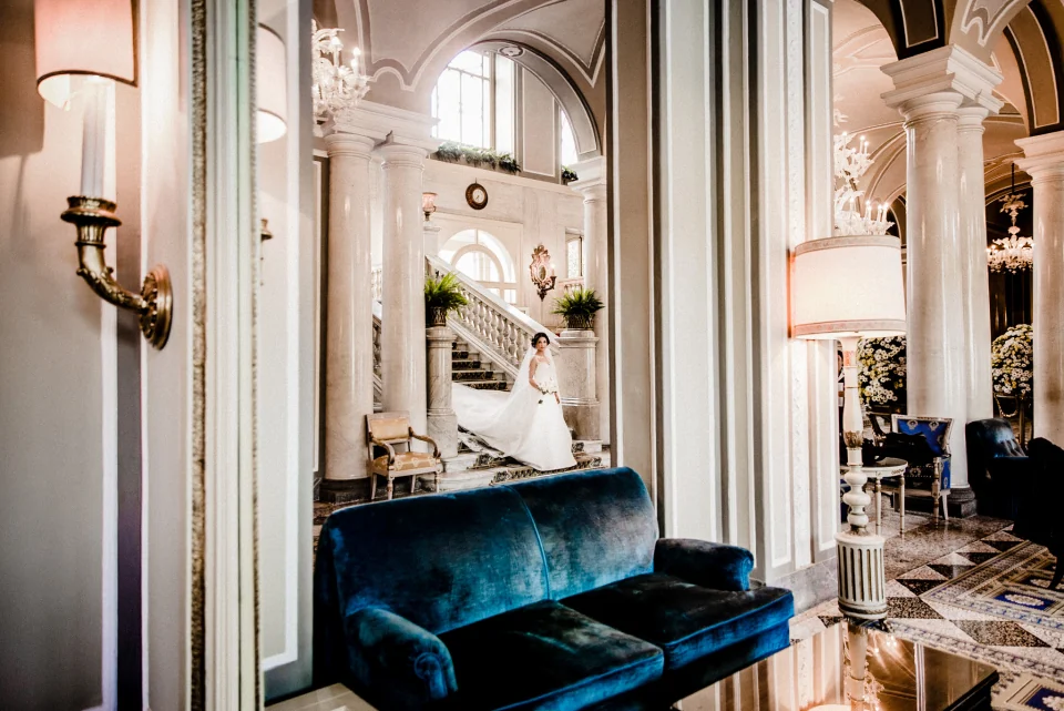A bride on a grand interior staircase, framed by pale arches and a blue velvet sofa.
